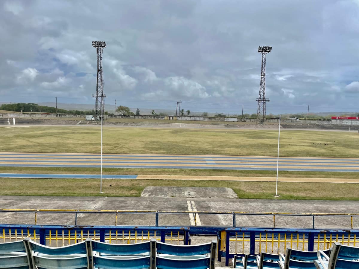 Campo de fútbol del Estadio Nacional de Barbados visto desde una grada, con pista azul y amarilla y torres de iluminación bajo un cielo nublado.