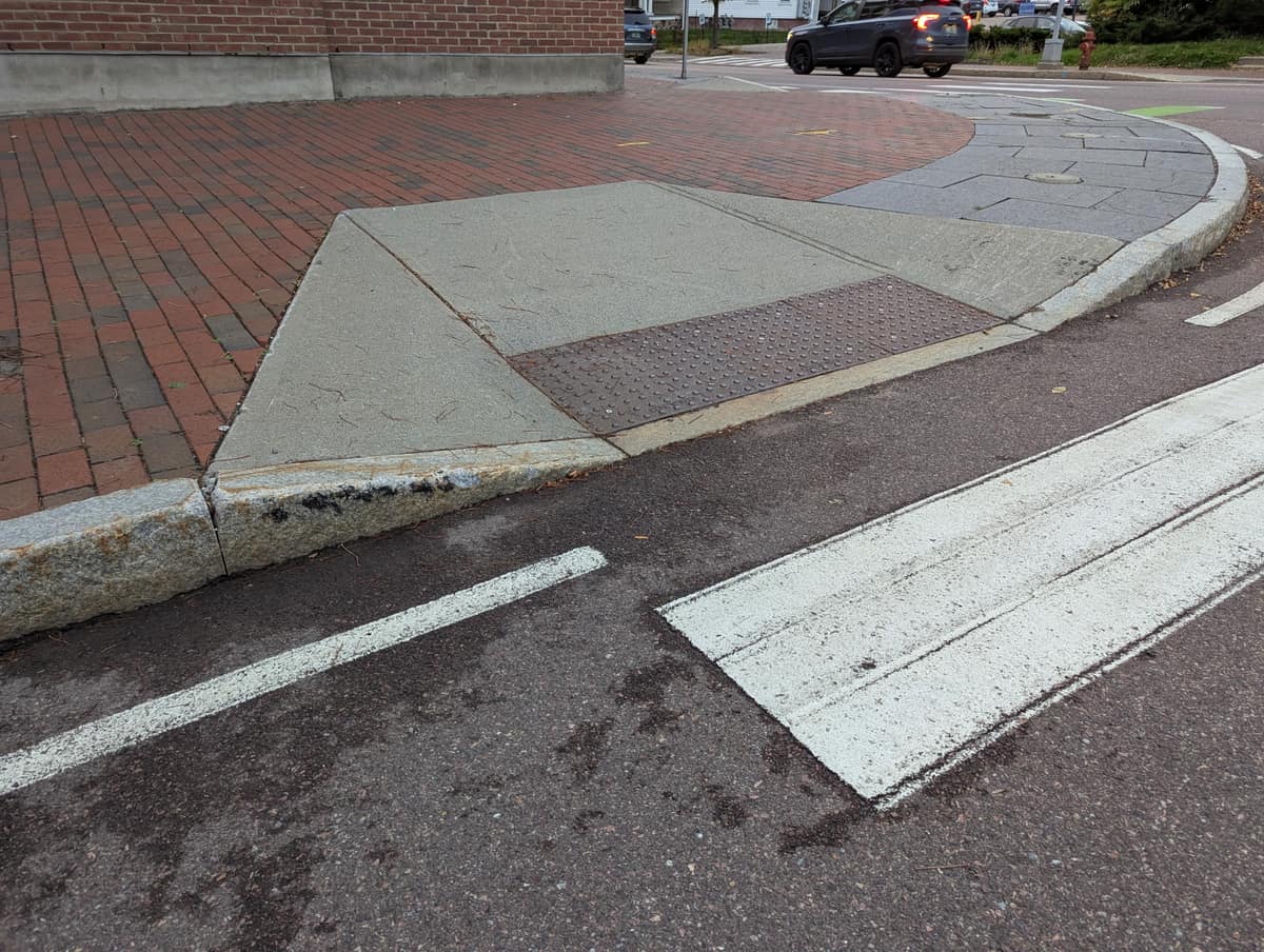 Acera con una rampa suave que desciende hasta la calzada y una franja rectangular de pavimento táctil junto al bordillo.