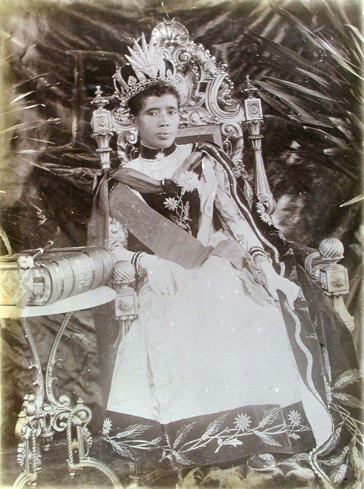 Retrato histórico de una soberana merina sentada en un trono, con corona, banda ceremonial y un gran libro sobre una mesa.