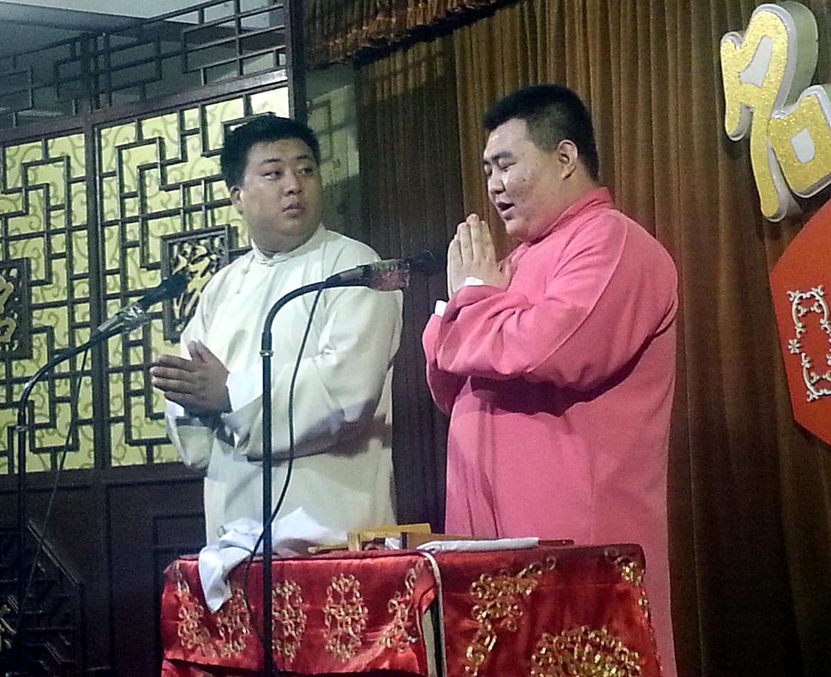 Dos intérpretes chinos con túnicas tradicionales conversan juntos sobre un escenario de xiangsheng en Tianjin.