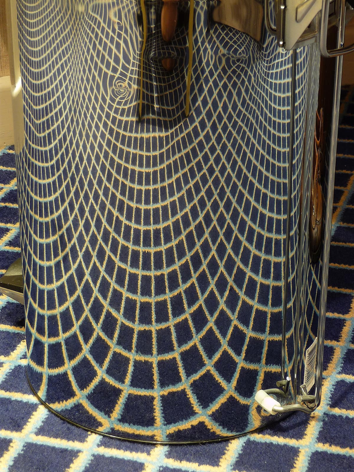 Alfombra con una figura deformada alrededor de un recipiente metálico cilíndrico que refleja una imagen reconocible.