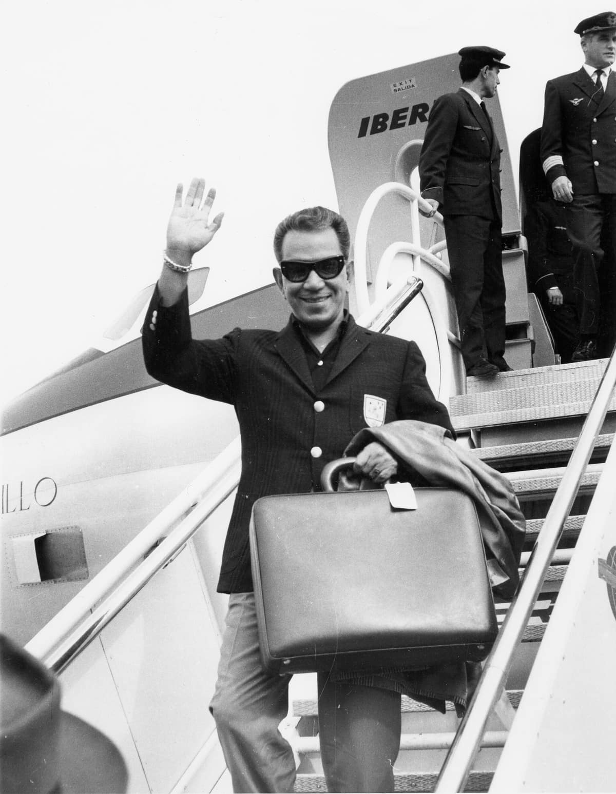Mario Moreno Cantinflas sonríe y saluda con una mano en una fotografía en blanco y negro tomada en 1964.